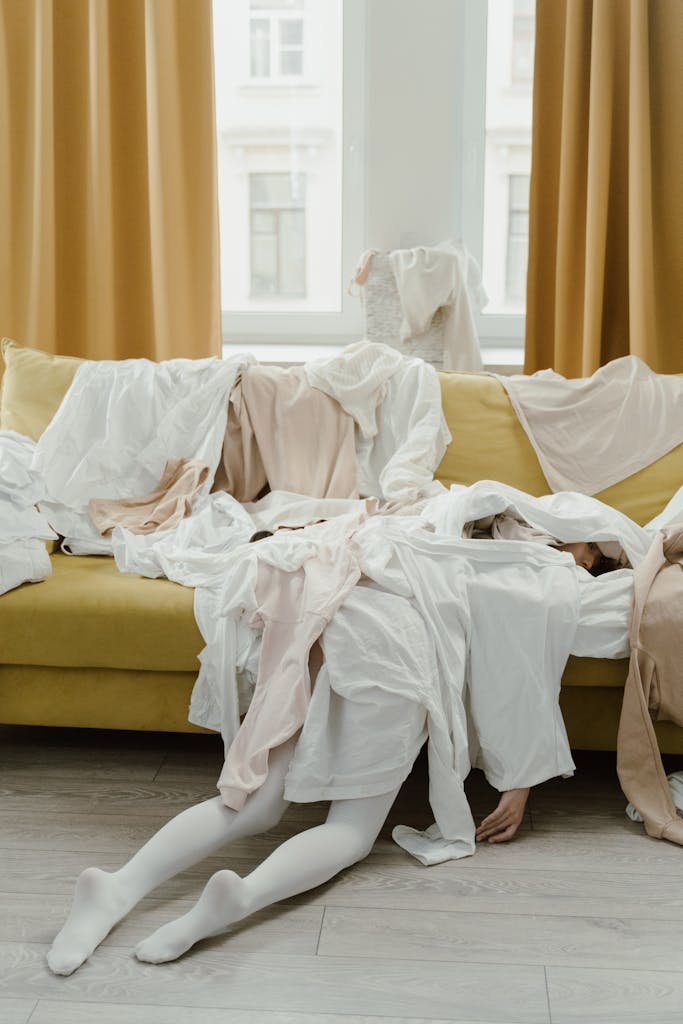 Woman buried under unorganized pile of clothes on a couch at home, expressing exhaustion.