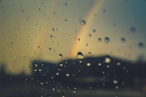 Close-up of raindrops on glass with a rainbow in the background, creating a moody atmospheric effect.