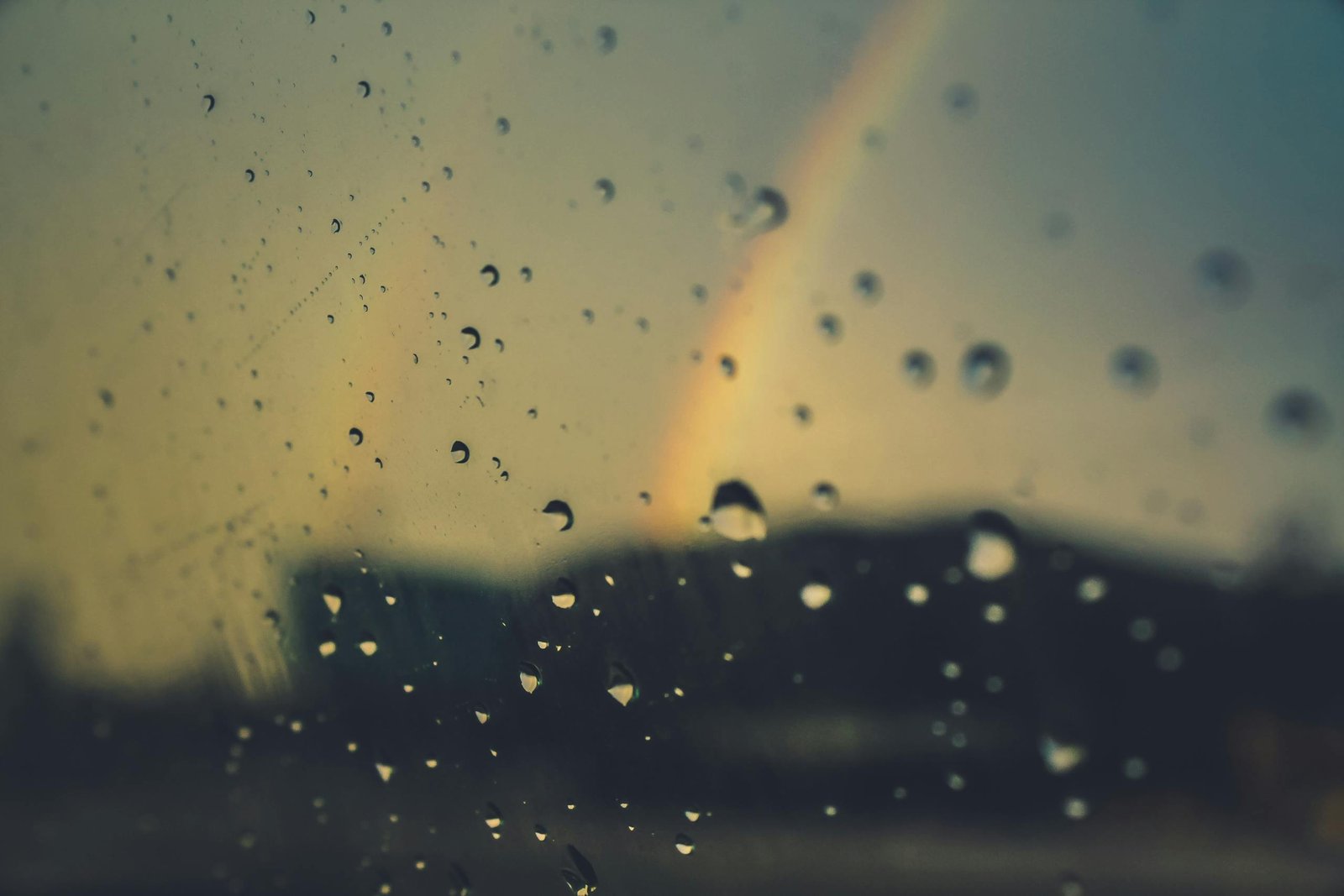 Close-up of raindrops on glass with a rainbow in the background, creating a moody atmospheric effect.